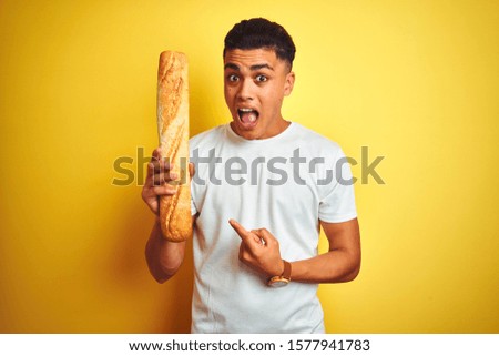 Young brazilian baker man holding bread standing over isolated yellow background very happy pointing with hand and finger