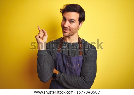 Young handsome shopkeeper man wearing apron standing over isolated yellow background with a big smile on face, pointing with hand and finger to the side looking at the camera.