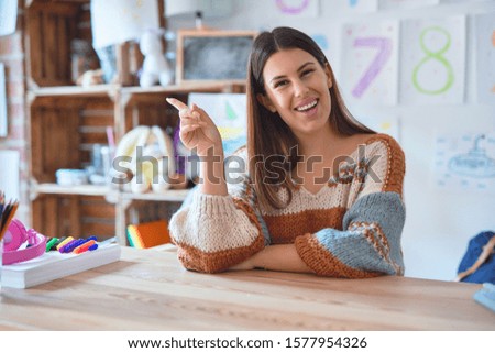 Young beautiful teacher woman wearing sweater and glasses sitting on desk at kindergarten with a big smile on face, pointing with hand and finger to the side looking at the camera.