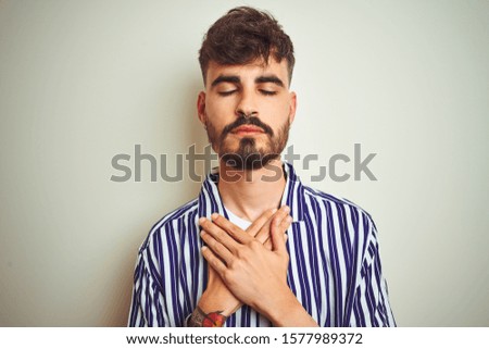Young man with tattoo wearing striped shirt standing over isolated white background smiling with hands on chest with closed eyes and grateful gesture on face. Health concept.