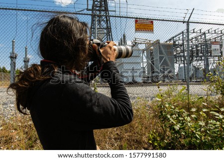 Back view of a man photographer with long hair, standing between plants and takes images of a high voltage electrical substation with a DSLR camera