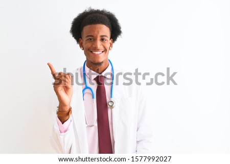 Young african american doctor man wearing sthetoscope over isolated white background with a big smile on face, pointing with hand and finger to the side looking at the camera.