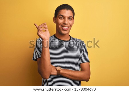Young handsome arab man wearing navy striped t-shirt over isolated yellow background with a big smile on face, pointing with hand and finger to the side looking at the camera.