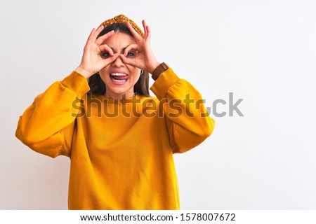 Young beautiful woman wearing yellow sweater and diadem over isolated white background doing ok gesture like binoculars sticking tongue out, eyes looking through fingers. Crazy expression.