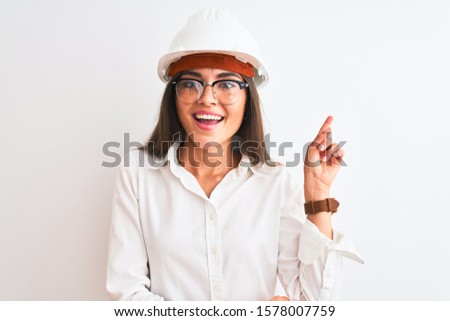 Young beautiful architect woman wearing helmet and glasses over isolated white background with a big smile on face, pointing with hand and finger to the side looking at the camera.