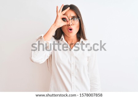 Young beautiful businesswoman wearing glasses standing over isolated white background doing ok gesture shocked with surprised face, eye looking through fingers. Unbelieving expression.