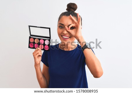 Young beautiful woman holding makeup standing over isolated white background with happy face smiling doing ok sign with hand on eye looking through fingers