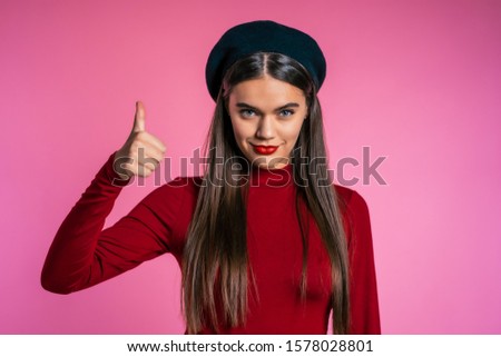 Winner. Success. Positive girl making thumbs up sign over pink background and smiles to camera. Body language. Young hipster woman with perfect red lips make-up and french hat.