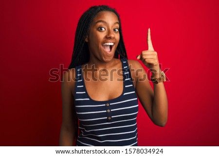 Young african american woman wearing striped t-shirt standing over isolated red background pointing finger up with successful idea. Exited and happy. Number one.