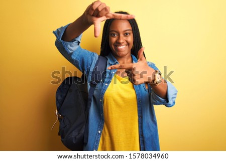 African american student woman wearing backpack standing over isolated yellow background smiling making frame with hands and fingers with happy face. Creativity and photography concept.