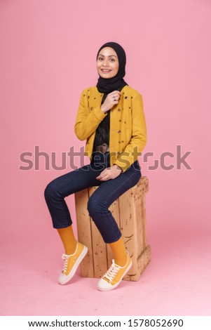 Fashionable hipster girl in dark blue jeans, long sleeves yellow leather jacket with hijab and sneakers isolated on pink background. Stylish Muslim hijab fashion lifestyle portraiture concept.