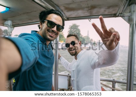 Young men sail on a ship and take a selfie against the background of a river in an Asian city. Tourists admire the views of the Asian city from a large boat and take pictures on the phone