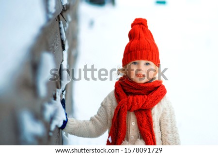 Funny little girl in a red knitted hat and scarf and white pullover playing outside in winter time. Kids play outdoors in winter. Children having fun at Christmas time. Winter fun.