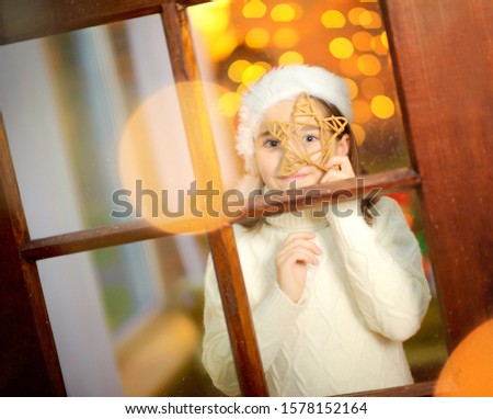 Funny girl in santa claus hat is played near the window, bright festive lights on the background. Christmas and New Year.