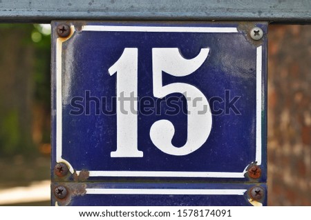 A blue house number plaque, showing the white coloured number fifteen (15)