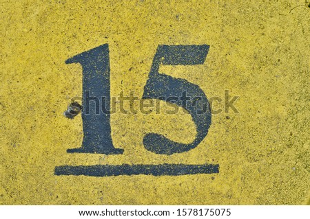 A painted house number on a yellow wall, showing the number fifteen (15)