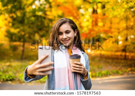 Happy woman holding takeaway coffee, taking a selfie on smart phone, outdoors in autumn.