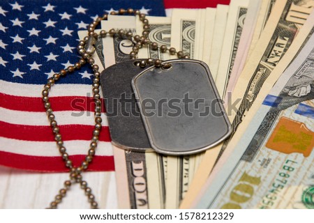 Against the background of the American flag and dollar bills lie army identification medallions. Concept: military pension, salary in the army, insurance for the military, mercenaries and contractors. Royalty-Free Stock Photo #1578212329