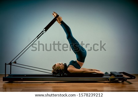 Young girl doing pilates exercises with a reformer bed. Beautiful slim fitness trainer on reformer gray background, low key, art light. Fitness concept Royalty-Free Stock Photo #1578239212