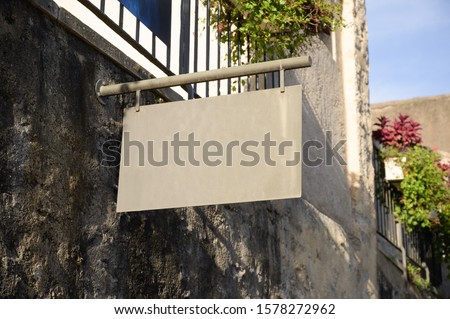A blank metal sign hinging from a stone wall with space for text.