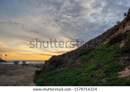 A Cloudy sunset on a beach in California with volleyball nets, lifeguard towers and people walking, seen from around the side of a mountain covered in green succulent ground cover and dried brush