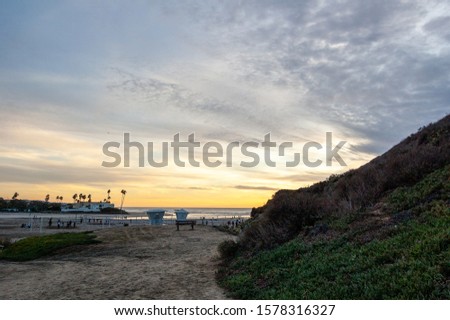 A Cloudy sunset on a beach in California with volleyball nets, lifeguard towers and people walking, seen from around the side of a mountain covered in green succulent ground cover and dried brush