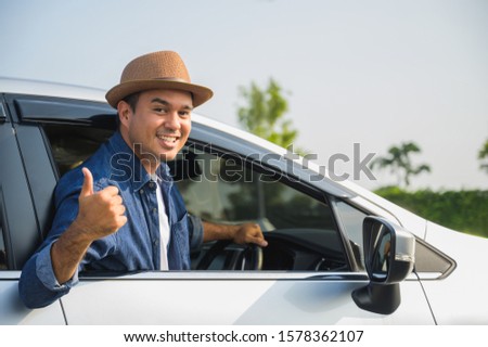 A traveler wearing a hat is driving a car and then pulling himself out of the car window. And thumbs up This picture is about a safe journey by car.