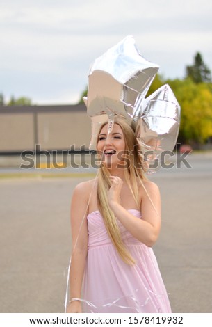 Pretty blonde girl holding silver star shaped balloons in a flowy white dress, outside on gravel in front of a blue and pink pastel colored wall. 