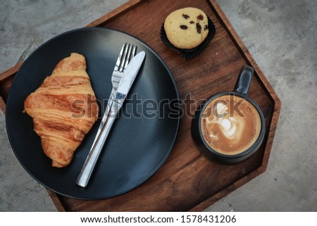 A cup of coffee with latte art on top and butter  croissant on tray, top view