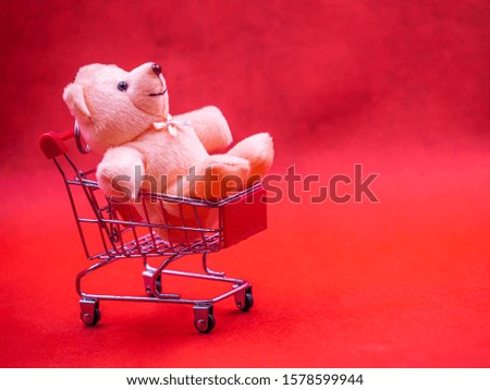 Closeup of cute brown teddy bear doll, sitting in trolley or supermarket cart with soft blurry vivid or vibrant gradient red texture and background for business, birthday concept.