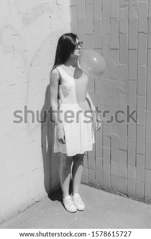 Black and white shot of Mellinial young brunette with rose cat sunglasses blowing pink bubble near rose wall