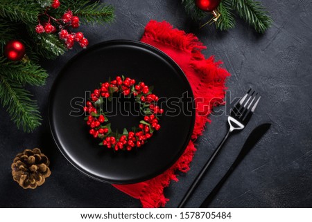 Christmas table setting with black craft plate and black cutlery with christmas decorations on black background. Top view with copy space.