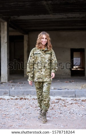 Young curly blond military woman, wearing ukrainian military uniform, posing for picture, smiling. Full-length portrait of soldier walking in front of ruined abandoned building, construction site.