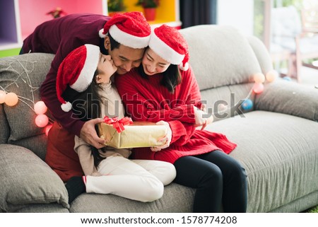 Young Asian family celebration in Christmas day, parent and daughter siting on sofa which hold give box, girl kiss father which smiling felling happy in living room at home morning time. Merry Xmas.