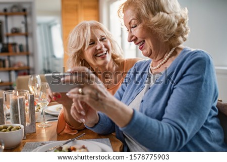 Happy senior women having fun while using smart phone and taking selfie at dining table. 