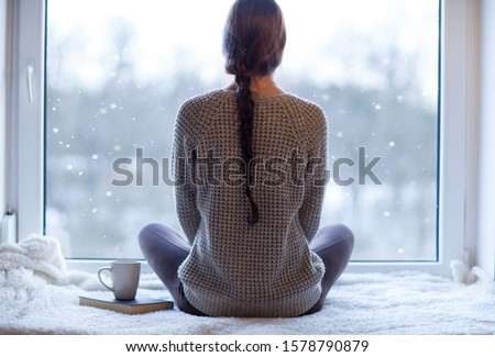 Thoughtful young brunette woman with book and cup of coffee looking through the window, blurry winter forrest landscape outside Royalty-Free Stock Photo #1578790879