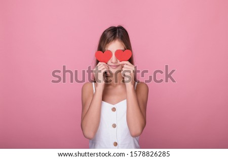 Cute young girl with long blond hair in a white dress holds two red paper hearts in her hands. Teen girl holding red hearts as glasses posing on a pink background. Valentine's day concept.