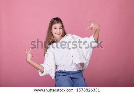 Funny cheerful young girl in a white jeans smiles broadly and looks straight, with her hands shows the sign of peace. The girl shows funny emotions on a pink background.