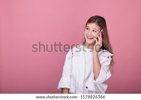 Beautiful young girl speaks on a pink phone and laughs. A girl in a white T-shirt and denim jacket holds the phone by her ear and laughs with a smile. Girl with a phone posing on a pink background.