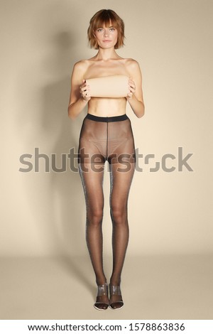 Full shot of a blonde lady in clear vinyl sandals and black sheer tights with silver lurex. The slim shirtless woman is holding an ivory white vase in front of her chest on the beige background. 