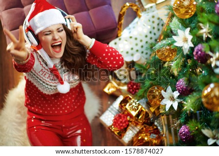 Smiling modern woman wearing red white Christmas sweater and elf santa hat listening to the music with headphones and showing victory gesture under decorated Christmas tree near present boxes.