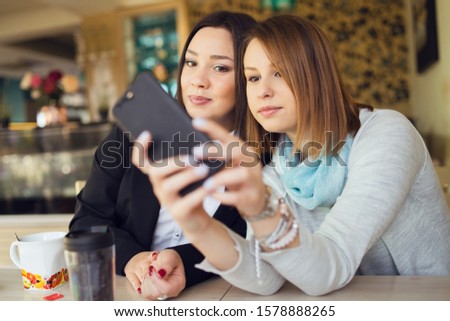Two young women female friends using smart phone smartphone at the restaurant cafe sitting by the table video chat call or taking selfie photos photographs
