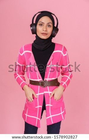 Fashionable hipster girl wearing jeans, bright long sleeves shirt with hijab and listening to music with wireless headphones isolated over pink background. Technology in a modern lifestyle concept.