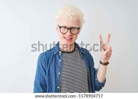 Young albino blond man wearing denim shirt and glasses over isolated white background showing and pointing up with fingers number two while smiling confident and happy.