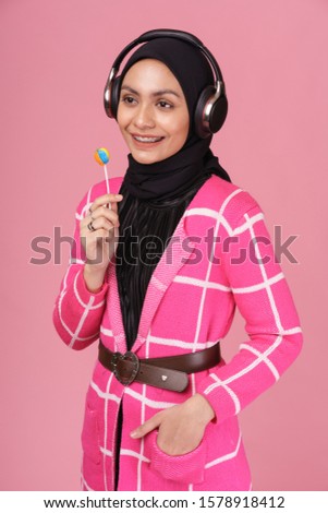 Cool young hipster girl wearing hijab, holding a sucking candy lolipop and listening to music with wireless headphones isolated over pink background. Technology in a modern lifestyle concept.