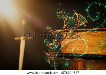 low key image of beautiful queen/king crown over antique book next to sword. fantasy medieval period. Selective focus. Glitter sparkle lights