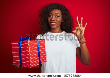 Young african american woman holding birthday gift standing over isolated red background doing ok sign with fingers, excellent symbol