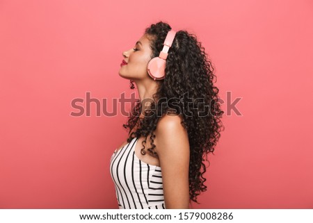 Image of happy african american woman with curly hair listening to music with headphones isolated over pink background