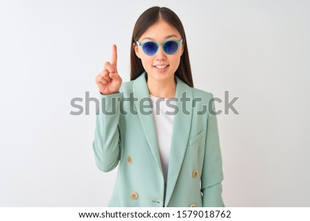 Chinese businesswoman wearing jacket and sunglasses over isolated white background surprised with an idea or question pointing finger with happy face, number one
