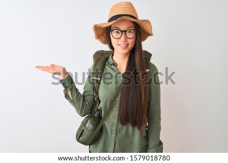 Chinese hiker woman wearing canteen hat glasses backpack over isolated white background smiling cheerful presenting and pointing with palm of hand looking at the camera.